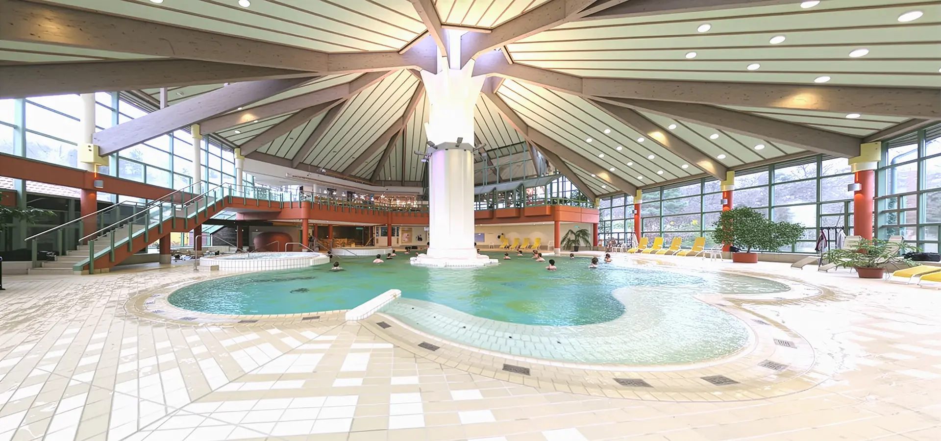 Innenansicht eines Schwimmbads mit rundem Becken, großer Säule in der Mitte und umlaufender Galerie mit Treppen.