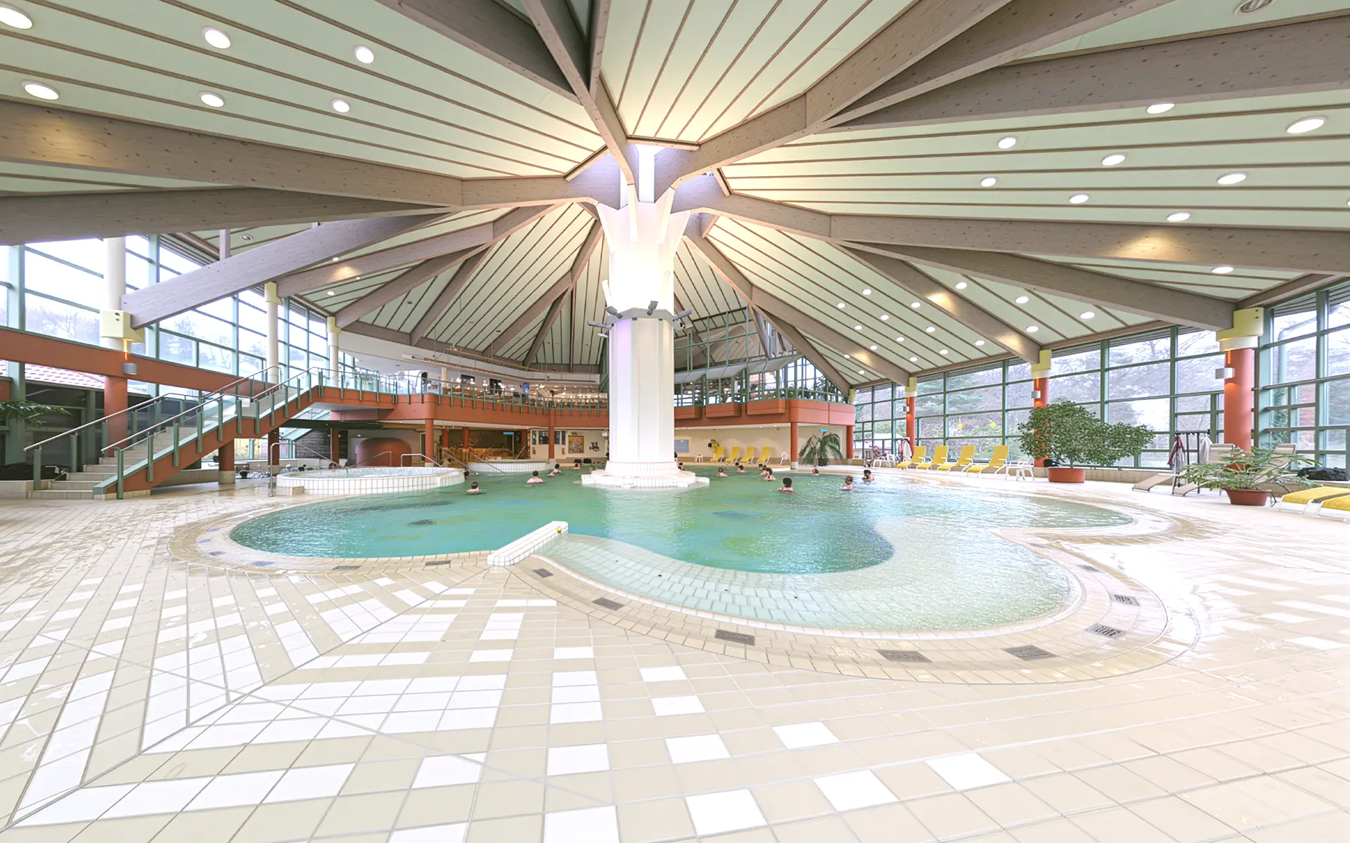 Innenansicht eines Schwimmbads mit rundem Becken, großer Säule in der Mitte und umlaufender Galerie mit Treppen.