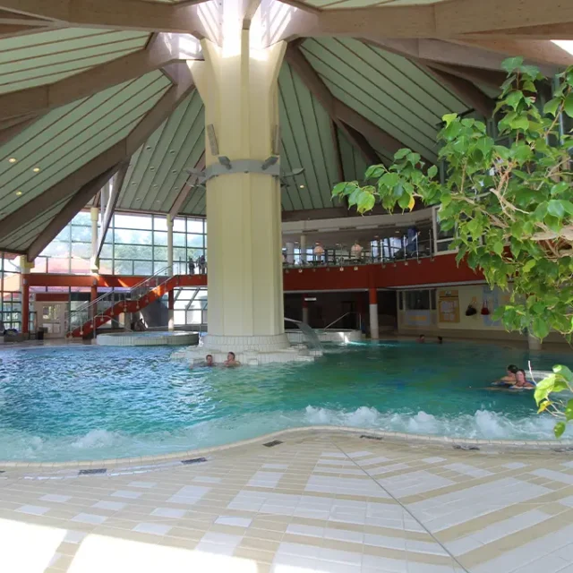 Innenansicht eines Schwimmbads mit rundem Becken, großer Säule in der Mitte und umlaufender Galerie mit Treppen.